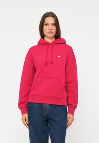 Rosa hettegenser laget av bomullsblanding, med en frontlomme, snorer i hetta, og en liten logo på brystet. Bært sammen med blå jeans.