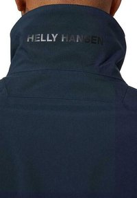 Marineblauwe jas met hoge kraag, met de merknaam "HELLY HANSEN" in zilver reflecterende tekst op de achterkant. Gladde textuur.
