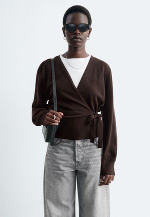 Femme portant un pull cache-cœur marron foncé, un haut blanc, un jean gris clair, des lunettes de soleil noires, un collier chaîne en argent et tenant un sac à épaule noir.
