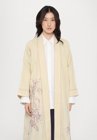 Bežový ľanový kabát v štýle kimono s ružovou vyšívanou kvetinovou výšivkou, ktorý má široké rukávy a prešívané okraje, navlečený na bielu košeľu.