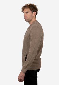 Cárdigan marrón claro con tejido texturizado, diseño ajustado y bolsillos laterales. Presenta un escote redondo y mangas largas.