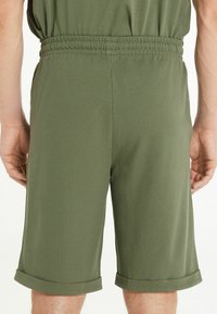 Pantalones cortos caquis fabricados en un tejido texturizado, con una cinturilla elástica y puños de dobladillo enrollados. El diseño es simple y funcional.