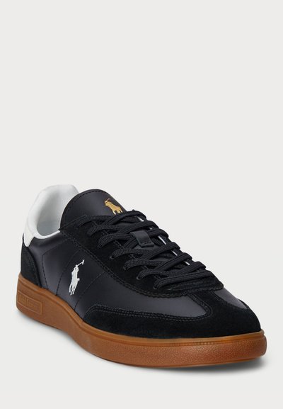 Melnās ādas un zamšādas sporta kurpes ar baltu apkaklīti, gumijas zoli, melnām šņorēm un baltiem un zelta logotipiem sānu daļā. Klasisks dizains un teksturēta apdare.