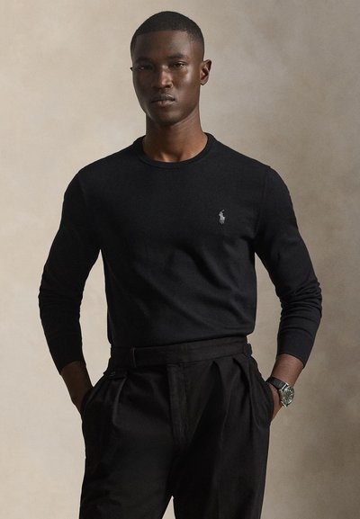 Mann mit kurzen Haaren, der ein schwarzes Langarmshirt und schwarze Hose trägt, steht mit den Händen in den Taschen vor einem neutralen Hintergrund.