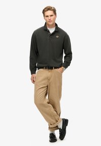 Black long-sleeve polo shirt with a collar, featuring a small logo; light beige loose-fit trousers; black shoes; white socks visible.