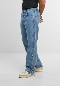 Hellblaue Jeans aus Denim mit geradem Schnitt, seitlichen Knopfdetails und kontrastierenden Nähten. Gestylt mit einem schwarzen Oberteil und Sneakers.