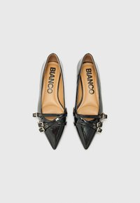 Scarpe lucide nere con punta a punta, dotate di cinturini alla caviglia e dettagli con fibbia, con una soletta beige contrassegnata con "BIANCO."