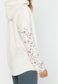 Vaalean beige huppari, jossa on kuviollinen kukkatesetti yhdellä hihalla. Pehmeä tekstuuri, rento malli ja yksinkertainen muotoilu.