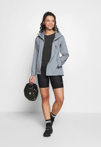 Femme portant une veste grise et un cuissard de cyclisme noir, tenant un casque de vélo noir, debout et souriante sur un fond blanc uni.