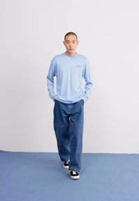 Camisa de manga longa azul clara com um pequeno logótipo, combinada com jeans de ganga escuros e folgados e ténis pretos e brancos.