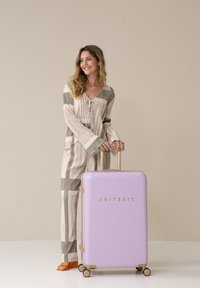 Harde koffer in pasteltint lavendel met gouden accenten, met een gladde textuur en vier wielen. Model staat ernaast in gestreepte kleding.