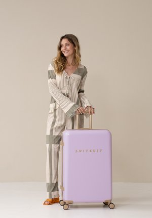 Hard-shell suitcase in pastel lavender with gold accents, featuring a smooth texture and four wheels. Model stands beside it wearing striped attire.