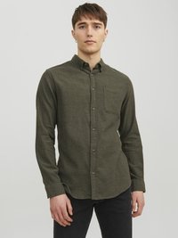Camisa de botones verde oliva, tela texturizada, mangas largas, bolsillo en el pecho, dobladillo redondeado, botones marrones, combinada con jeans oscuros.
