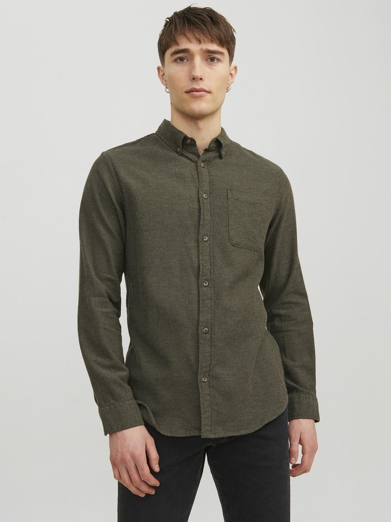 Camisa de botones verde oliva, tela texturizada, mangas largas, bolsillo en el pecho, dobladillo redondeado, botones marrones, combinada con jeans oscuros.
