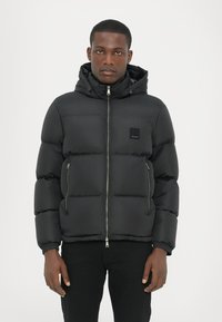 Schwarze gepolsterte Jacke mit einem Reißverschluss vorne, Kapuze und elastischen Bündchen. Verfügt über seitliche Reißverschlusstaschen und ein kleines Markenlabel auf der Brust.