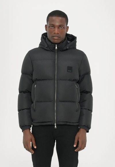 Veste rembourrée noire avec une fermeture éclair à l'avant, une capuche et des poignets élastiques. Comprend des poches latérales zippées et une petite étiquette de marque sur la poitrine.