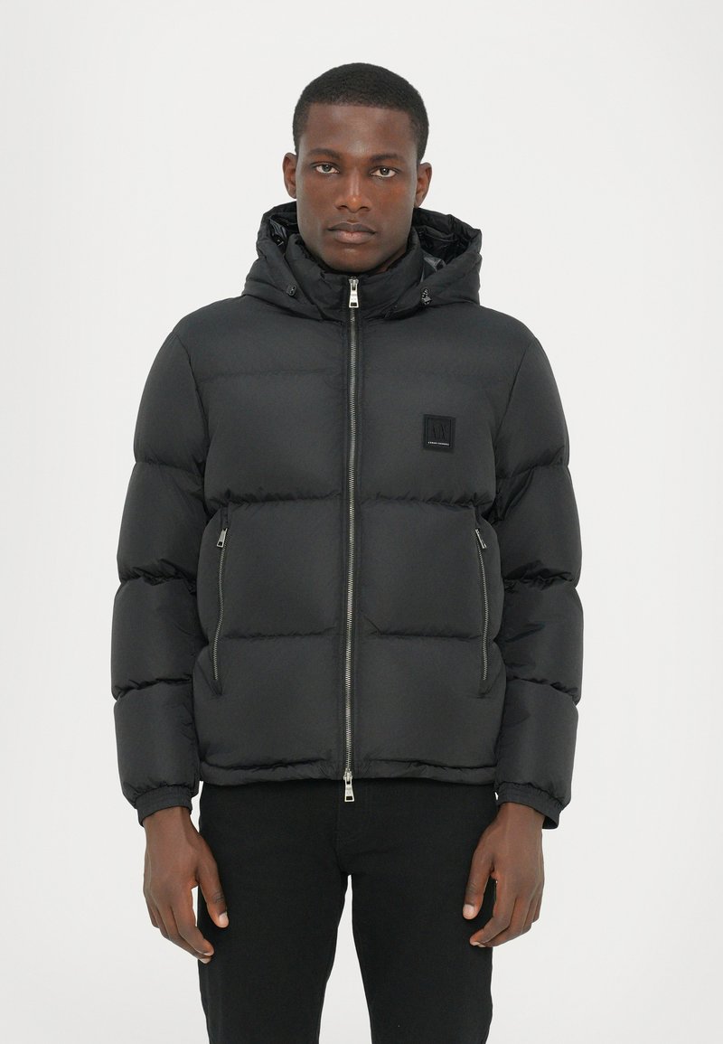 Schwarze gepolsterte Jacke mit einem Reißverschluss vorne, Kapuze und elastischen Bündchen. Verfügt über seitliche Reißverschlusstaschen und ein kleines Markenlabel auf der Brust.