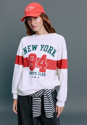 Jeune femme portant un sweat-shirt blanc avec le texte New York 94 Sports Club en rouge et vert, casquette rouge et vêtement rayé noué autour de la taille.