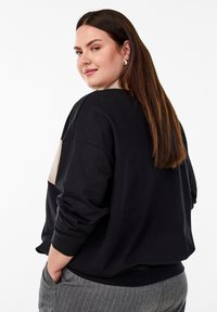 Svart sweatshirt med oversized passform, kontrasterande beige panel på baksidan, ribbade muddar och strukturerad tyg. Modellen har långt brunt hår.
