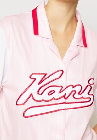 Różowa koszula w paski z czerwonym kołnierzykiem, z dużym czerwonym haftowanym napisem "Kani." Materiał wydaje się mieć teksturę, z detalami guzików.