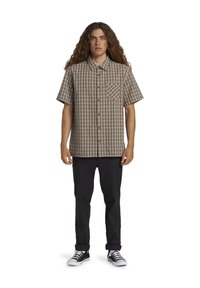 Chemise à manches courtes avec un motif à carreaux brun et blanc, dotée d'une poche poitrine et d'une coupe décontractée, associée à un pantalon noir.
