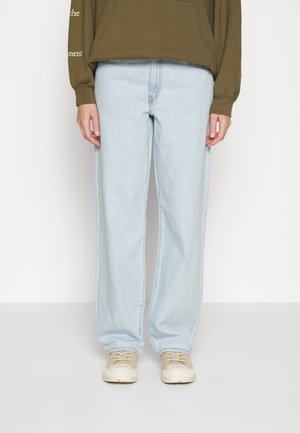 Person trägt hellblaue Jeans mit geradem Bein, beige Turnschuhe und ein olivgrünes Sweatshirt mit Tasche und Text auf dem Ärmel.