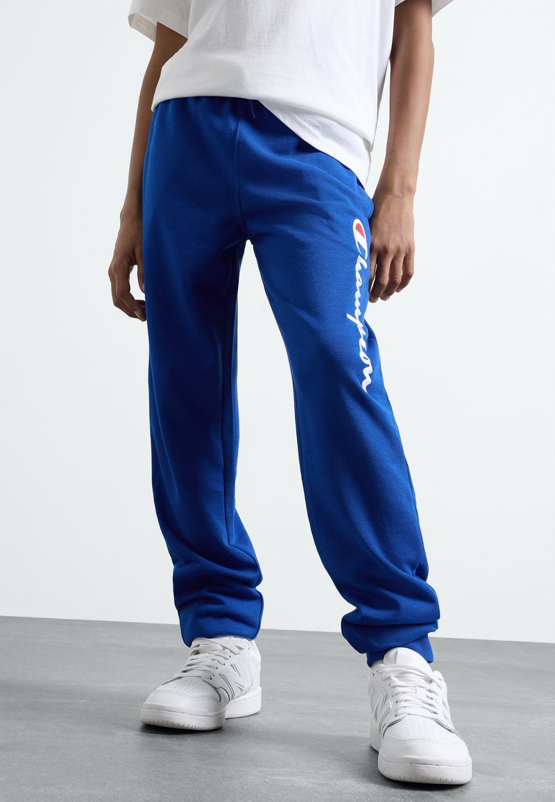 Blue sweatpants with elastic waistband, featuring a white "Champion" logo along the side. Paired with white sneakers on a gray floor.