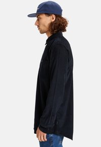 Navy corduroy shirt with long sleeves, a chest pocket, and side slits. Features textured fabric and a relaxed fit.