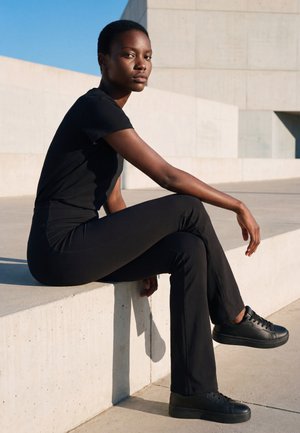 Une femme vêtue de noir et chaussée de baskets est assise sur un rebord en béton à l’extérieur et regarde en direction de l’appareil photo sous un soleil éclatant.