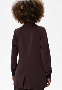 Donkerbruine lange mouwen jas met een reverskraag, voorzien van een middenachternaad en zijsplitten voor extra bewegingsvrijheid. Zachte textuur.