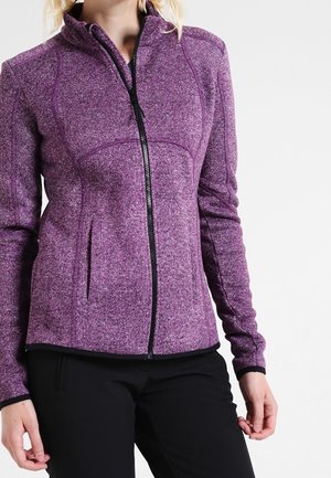 Femme portant une veste polaire violette zippée et un pantalon noir, debout, les mains détendues le long du corps.