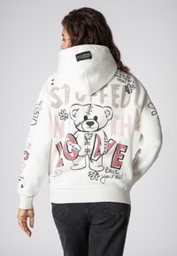White hoodie with a large graphic of a stitched teddy bear, accompanied by text and floral accents in pink and black. Features a hood and ribbed cuffs.