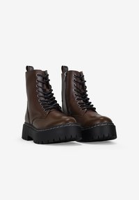 Par de botas de cuero marrón oscuro con cordones, suelas de plataforma negra gruesa y cremalleras laterales sobre fondo blanco.