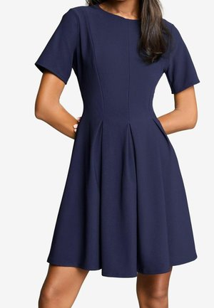 Vrouw in een marineblauwe, korte mouwen, knielange jurk met een aansluitende taille en geplooide rok, handen op haar heupen.