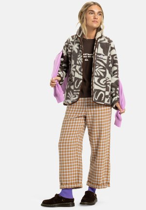 Veste en polaire avec un motif floral brun et crème, associée à un t-shirt graphique marron, un pantalon à carreaux et des chaussettes violettes.