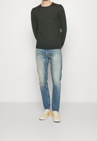 Maglione a maniche lunghe verde scuro, jeans azzurri chiari e sneaker crema con accenti gialli. Outfit casual con design slim fit.