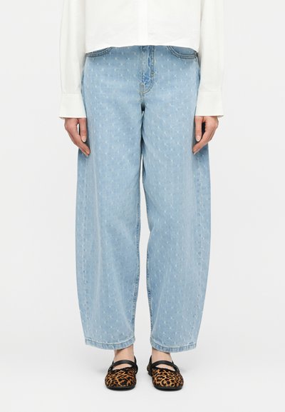 Φαρδιά τζιν παντελόνια σε ανοιχτό μπλε με διακριτικό μοτίβο αστεριών, σε συνδυασμό με λευκή μπλούζα με μακριά μανίκια και παπούτσια Mary Jane με λεοπάρ σχέδιο, σε μοντέλο.