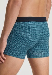 Boxershorts in donker teal met een geometrisch patroon. Het materiaal lijkt glad; de tailleband is effen zwart met de merknaam zichtbaar.