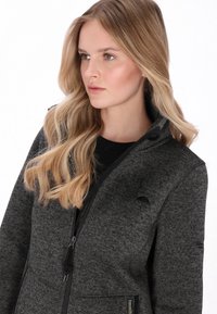 Szőrme pulóver szürke színben, elöl cipzárral, magas gallérral és finom textúrával. Fekete díszítésekkel és varratokkal kontrasztot teremt.