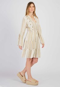 Weißes Kleid mit goldenen Mustern, gestuftem Design und langen Glockenärmeln. Kombiniert mit beigen Plateauschuhen zum Hineinschlüpfen.