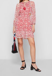 Vestito floreale rosso con maniche lunghe, dettaglio in pizzo, inserti laterali e fiocco con nappe. Abbinato a sandali neri con tacco a blocco e piccola borsa.