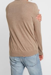 Hellbrauner Strickpullover mit rundem Halsausschnitt, gerippten Bündchen und Saum. Getragen über blauen Denim-Jeans, die eine lockere Passform zeigen.