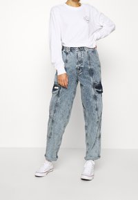 Camicia bianca a maniche lunghe con una piccola grafica sul petto abbinata a pantaloni cargo in denim azzurro oversize e sneakers bianche.