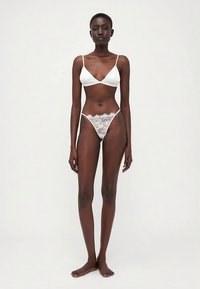 Hvit blonde bralette med tynne stropper, parret med hvit blonde tanga med bølgete kanter og blomstermønster. Modellen står barbeint.