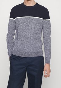 Pull en tricot marine et gris chiné avec un col côtelé. Présente une bande horizontale blanche. Fini texturé, poignets ajustés.
