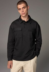 Sort langærmet polo shirt lavet af tekstureret stof med krave og to brystlommer med knaplukninger.