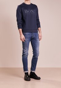 Námořnická modrá mikina se znakem "BOSS HUGO BOSS" v matném provedení, v kombinaci s přiléhavými modrými džíny a černými botami. Jednoduchý, ležérní design.