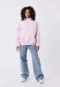 Lichtroze pullover sweatshirt met een kwart rits, een voorkant pocket, witte piping en een elastische zoom, gecombineerd met losse lichtblauwe jeans.