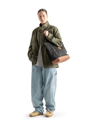Black and brown tote bag made of durable fabric, featuring a logo patch and a structured shape, worn over a light gray shirt and baggy jeans.