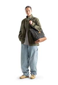 Svart tygväska med brun botten och en sydd logotypmärke, bärs av en person i en olivgrön jacka och vida blå jeans.